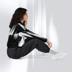 A woman sits on a white floor against a white wall, wearing a black and white striped hooded jacket, black jogger pants, white socks, and white sneakers.