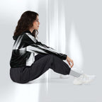 A woman sits on a white floor against a white wall, wearing a black and white striped hooded jacket, black jogger pants, white socks, and white sneakers.