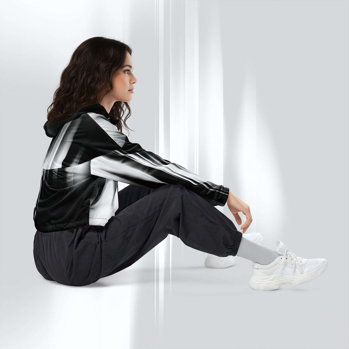 A woman sits on a white floor against a white wall, wearing a black and white striped hooded jacket, black jogger pants, white socks, and white sneakers.