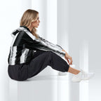 A woman sits on a glossy white floor in a minimalist studio setting, wearing a black and white striped hooded jacket, black jogger pants, and white sneakers.