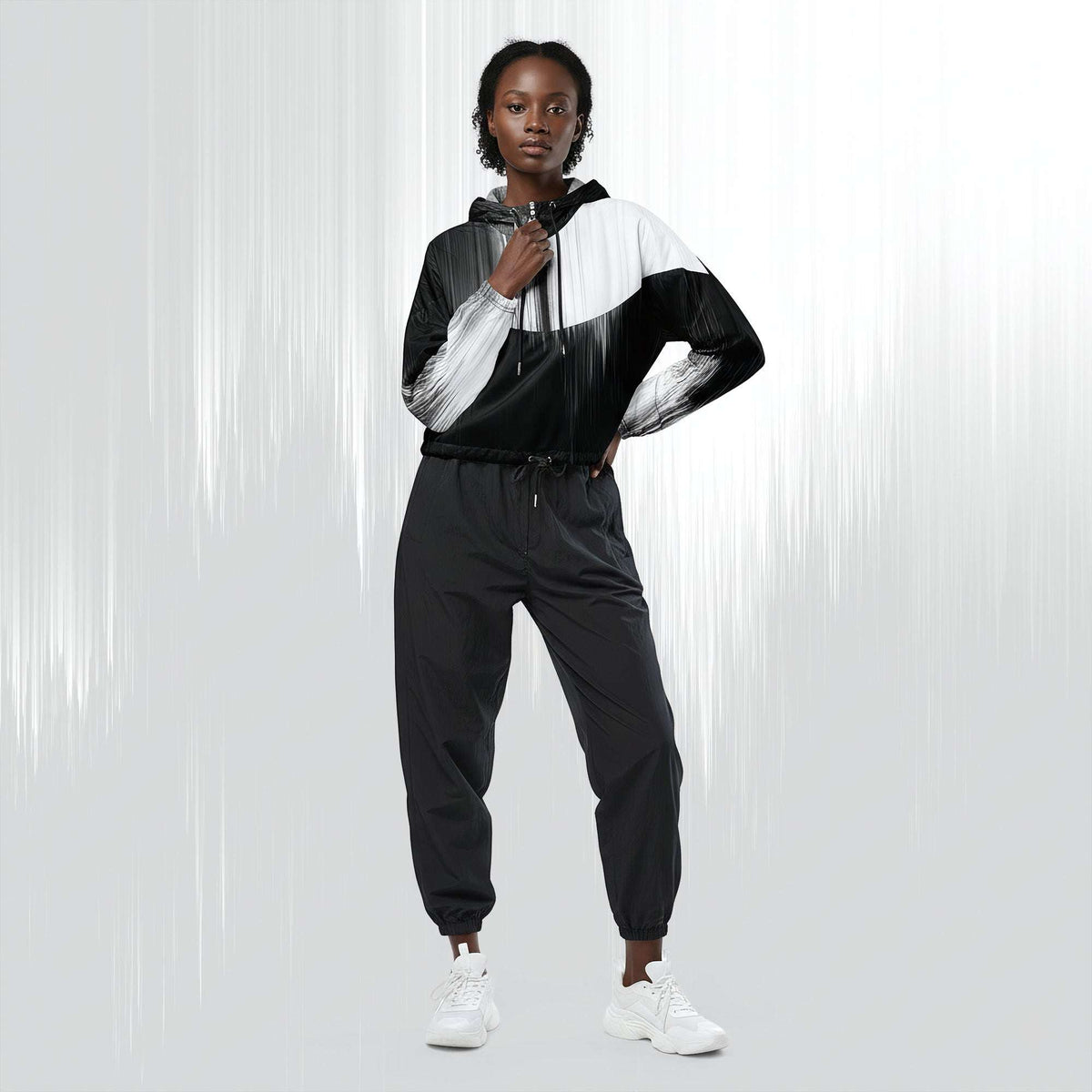 A model wearing a black and white color-blocked zip-up hoodie with matching black jogger pants and white sneakers, standing against a light gray studio background with vertical light streaks.