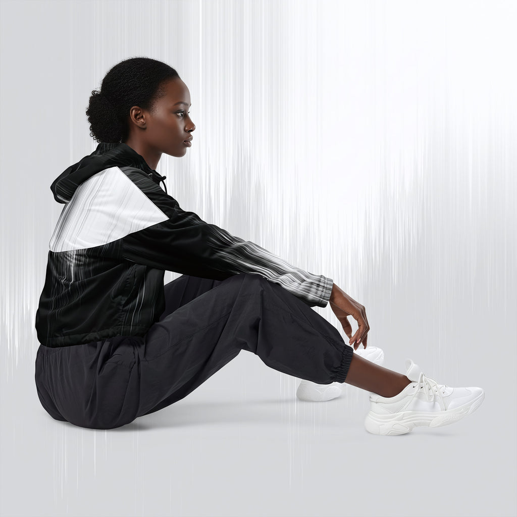 A model sits on the floor wearing a black and white windbreaker jacket, black jogger pants, and white sneakers against a light gray studio background with vertical light streaks.