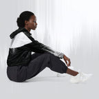 A model sits on the floor wearing a black and white windbreaker jacket, black jogger pants, and white sneakers against a light gray studio background with vertical light streaks.