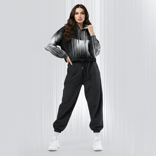 A woman modeling a black and white striped zip-up hoodie with black jogger sweatpants and white sneakers, standing against a light gray textured studio background.