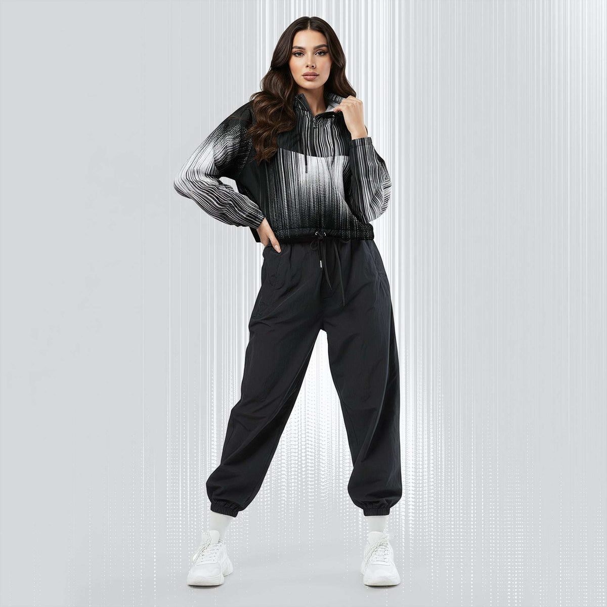A woman modeling a black and white striped zip-up hoodie with black jogger sweatpants and white sneakers, standing against a light gray textured studio background.