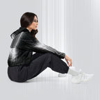 A woman sits on the floor in a studio setting, wearing a black hooded jacket with a white-to-gray gradient pattern on the sleeves and upper body, black jogger pants, and white sneakers.