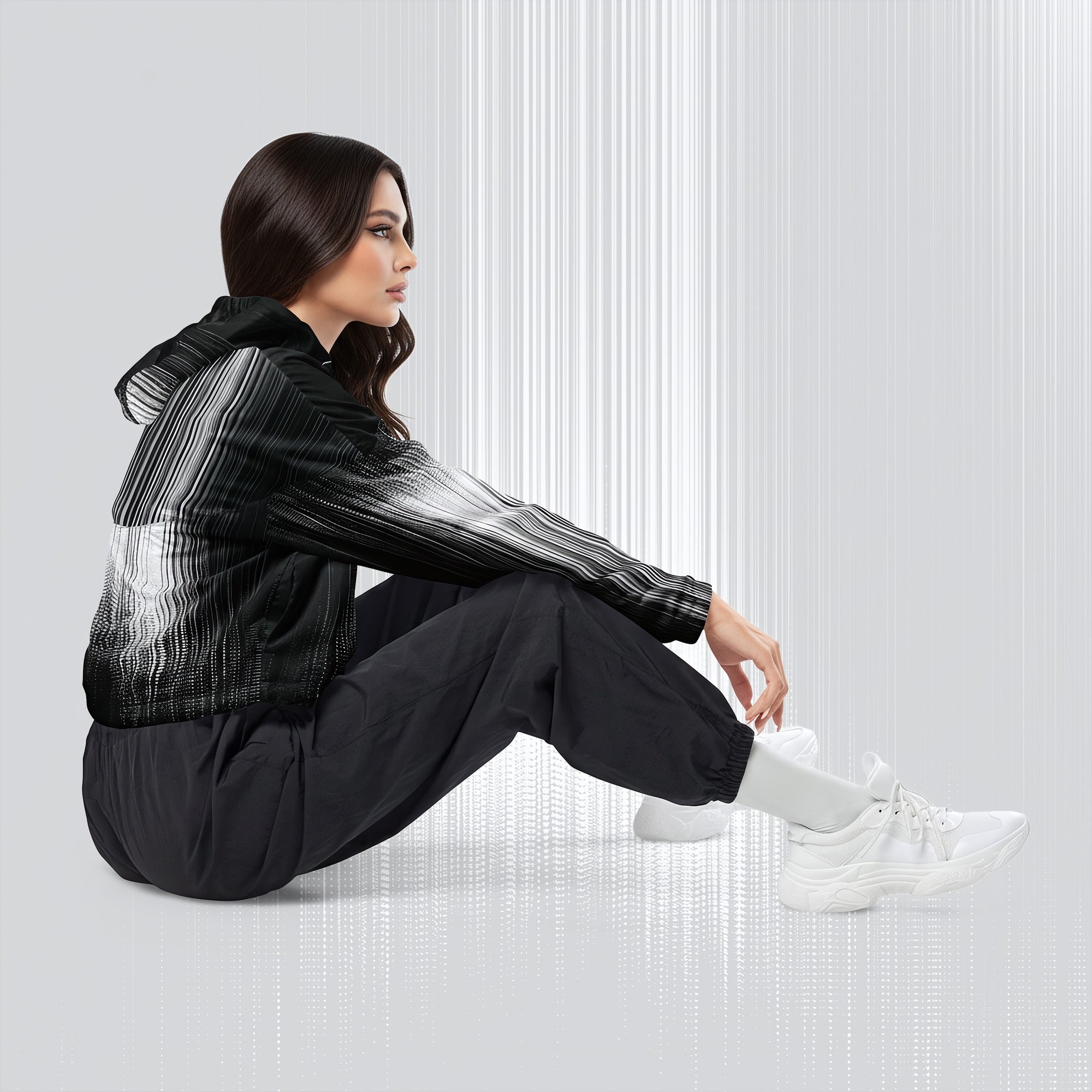A woman sits on the floor in a studio setting, wearing a black hooded jacket with a white-to-gray gradient pattern on the sleeves and upper body, black jogger pants, and white sneakers.