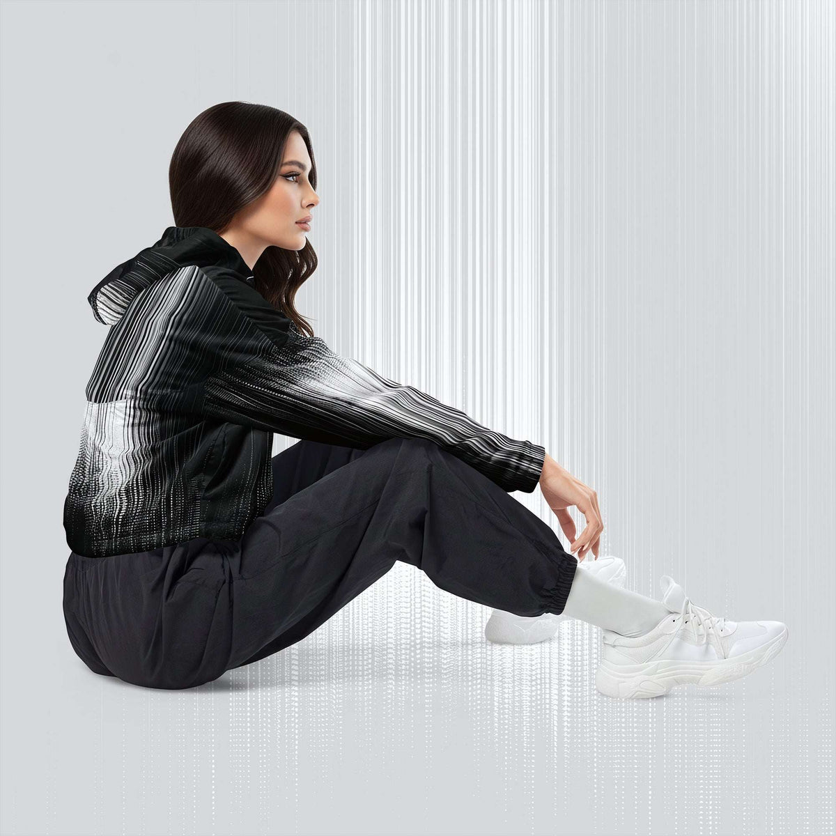 A woman sits on the floor in a studio setting, wearing a black hooded jacket with a white-to-gray gradient pattern on the sleeves and upper body, black jogger pants, and white sneakers.