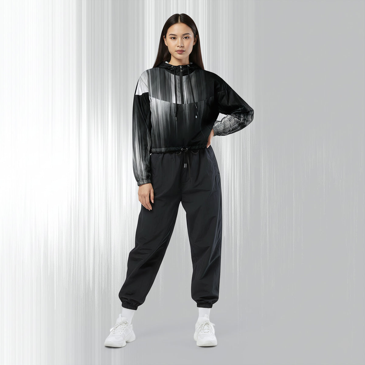 A woman modeling a black and gray gradient patterned hooded windbreaker jacket with matching black jogger pants and white sneakers, set against a light gray studio background with vertical light streaks.