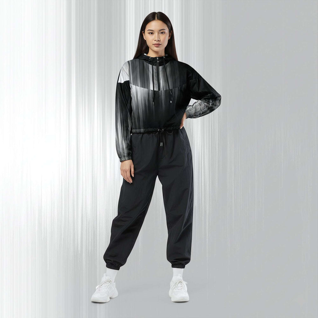 A woman modeling a black and gray gradient patterned hooded windbreaker jacket with matching black jogger pants and white sneakers, set against a light gray studio background with vertical light streaks.