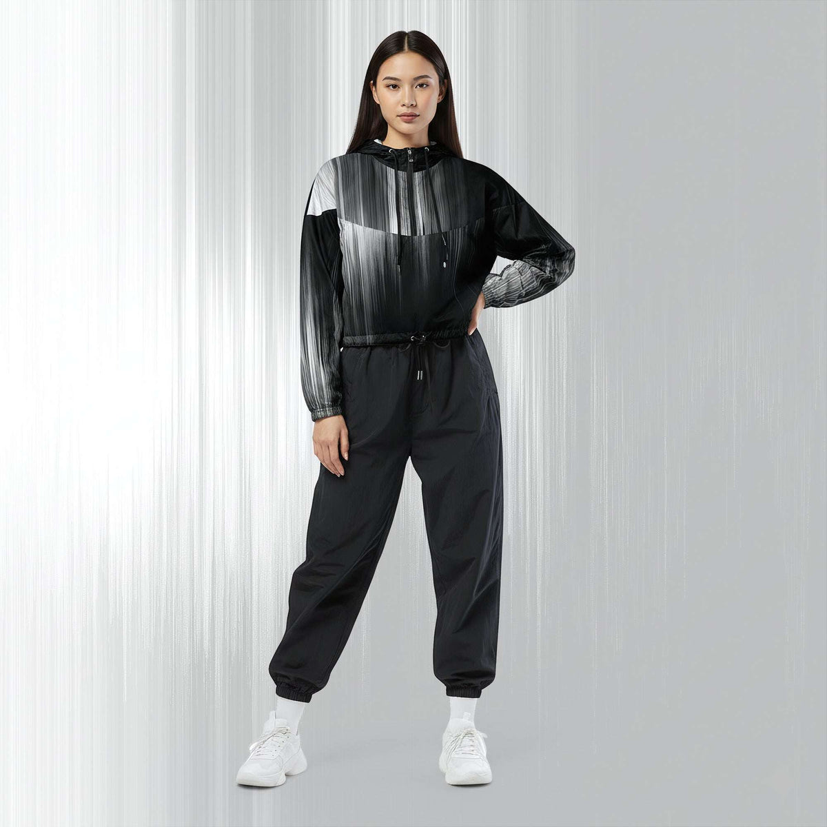 A woman modeling a black and gray gradient patterned hooded windbreaker jacket with matching black jogger pants and white sneakers, set against a light gray studio background with vertical light streaks.