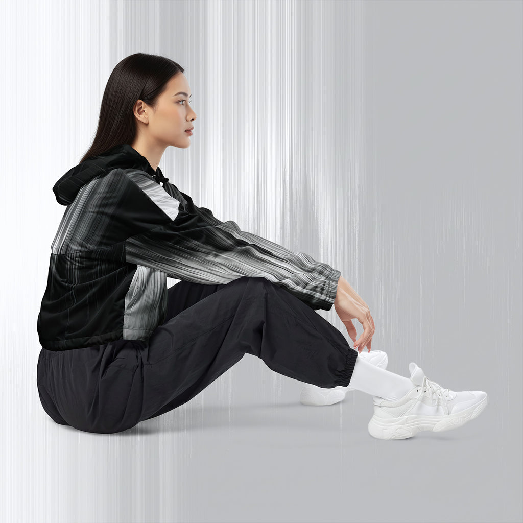 A woman sits on the floor wearing a black and gray patterned hooded jacket, black jogger pants, and white sneakers against a light gray studio background.