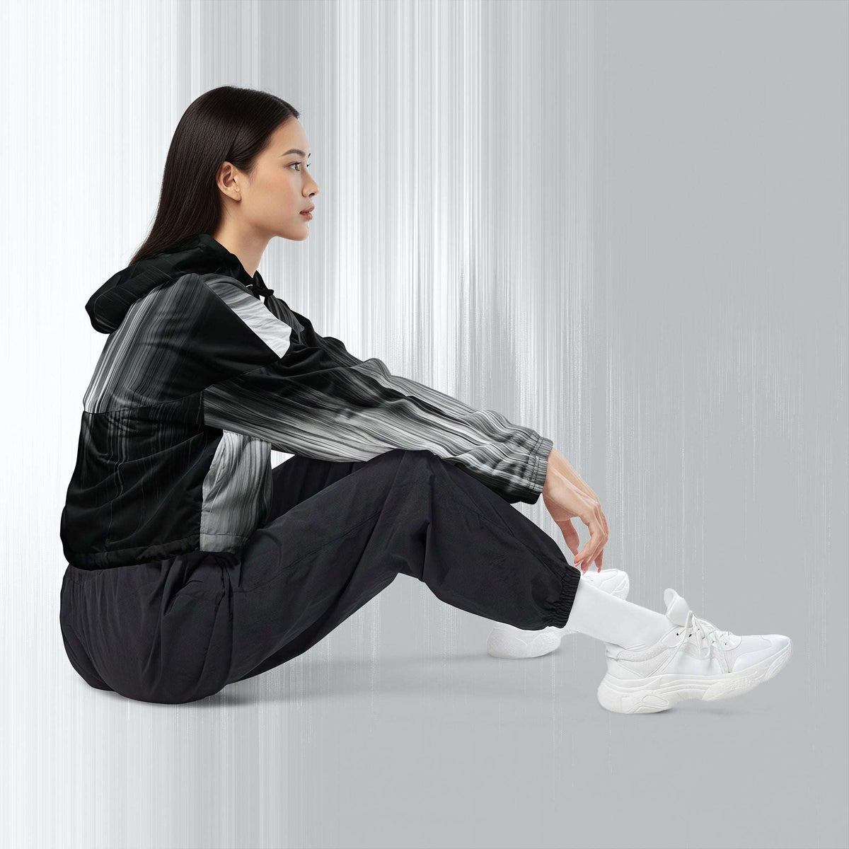 A woman sits on the floor wearing a black and gray patterned hooded jacket, black jogger pants, and white sneakers against a light gray studio background.