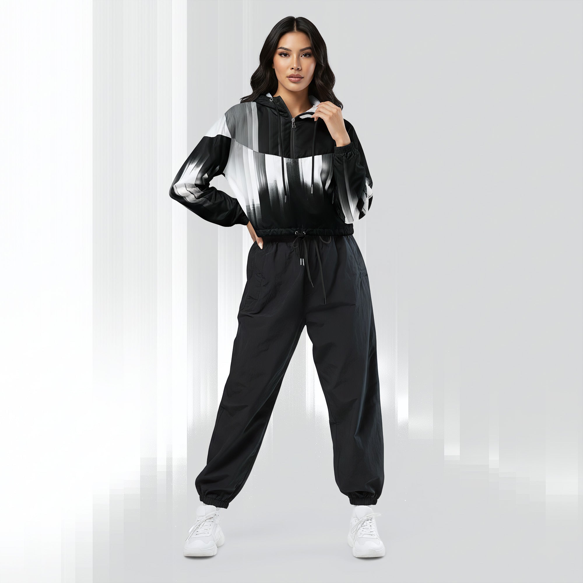 A woman modeling a black and white abstract-patterned hoodie with black jogger pants and white sneakers, standing against a light gray studio background.