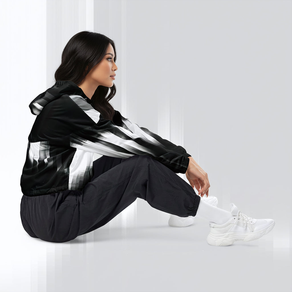 A woman sits on a glossy white floor wearing a black windbreaker jacket with bold white and gray brushstroke patterns, black jogger pants, and white sneakers.