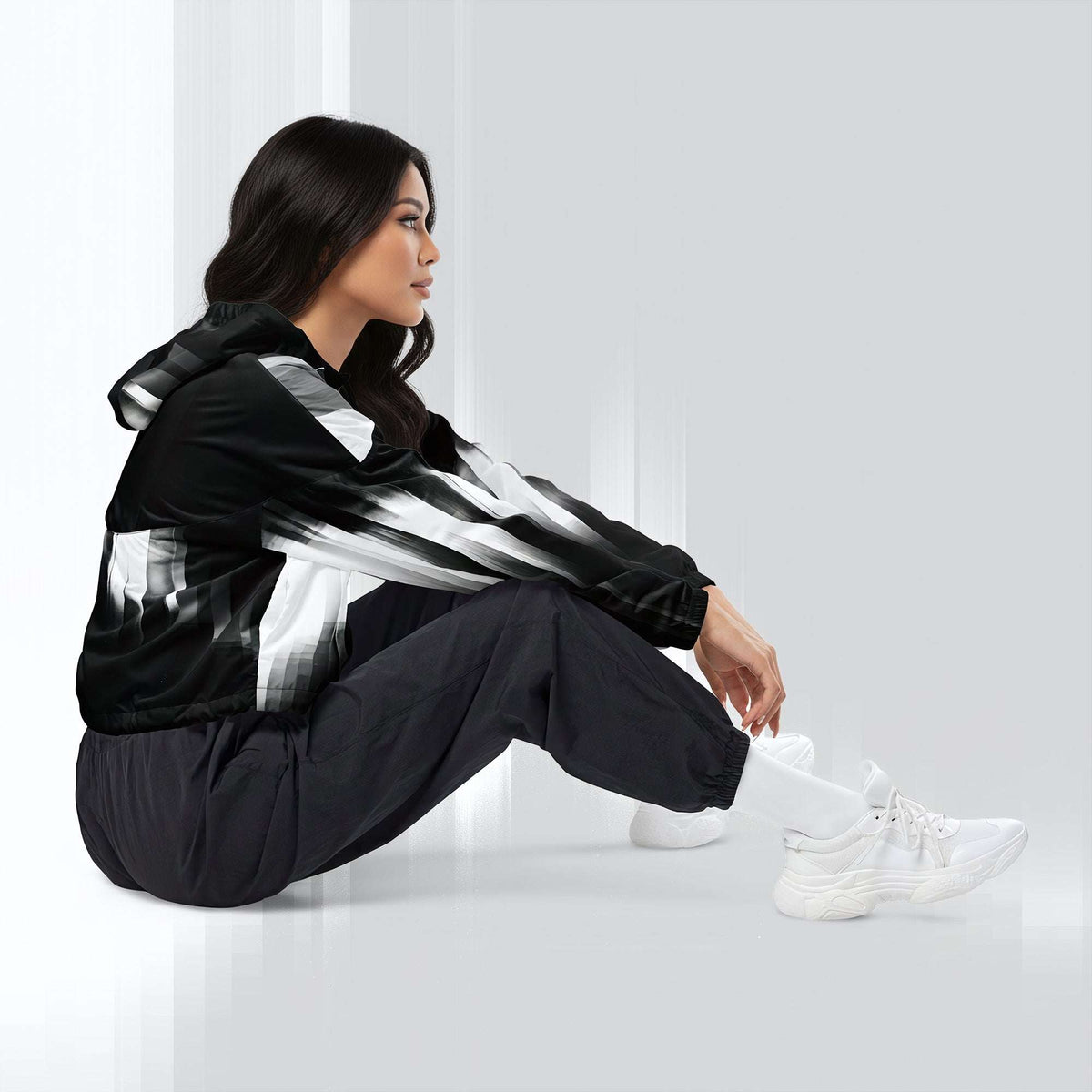 A woman sits on a glossy white floor wearing a black windbreaker jacket with bold white and gray brushstroke patterns, black jogger pants, and white sneakers.
