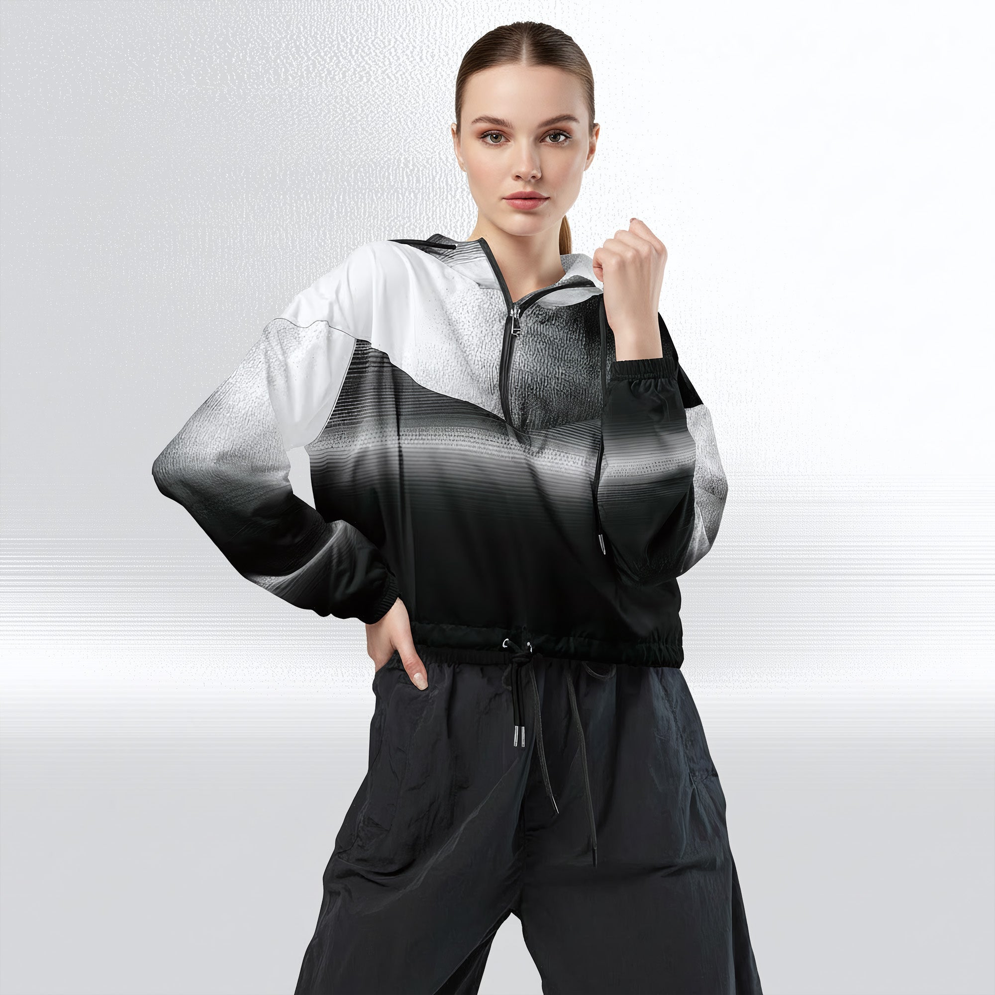 Female model wearing the HER Cropped Dither Distortion Windbreaker, three‑quarter front view. The cropped windbreaker has a black‑to‑white dither gradient pattern, long sleeves and a half‑zip front. The model holds the collar with one hand, the hood is down, and the jacket is paired with black trousers against a light grey background