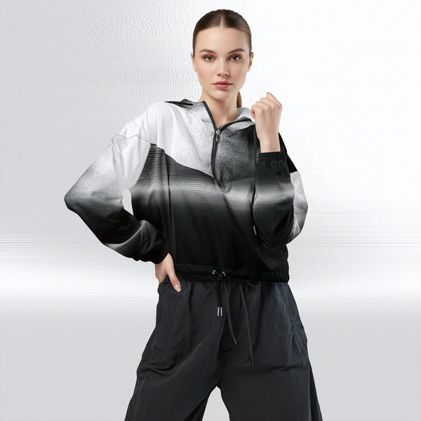 Female model wearing the HER Cropped Dither Distortion Windbreaker, three‑quarter front view. The cropped windbreaker has a black‑to‑white dither gradient pattern, long sleeves and a half‑zip front. The model holds the collar with one hand, the hood is down, and the jacket is paired with black trousers against a light grey background