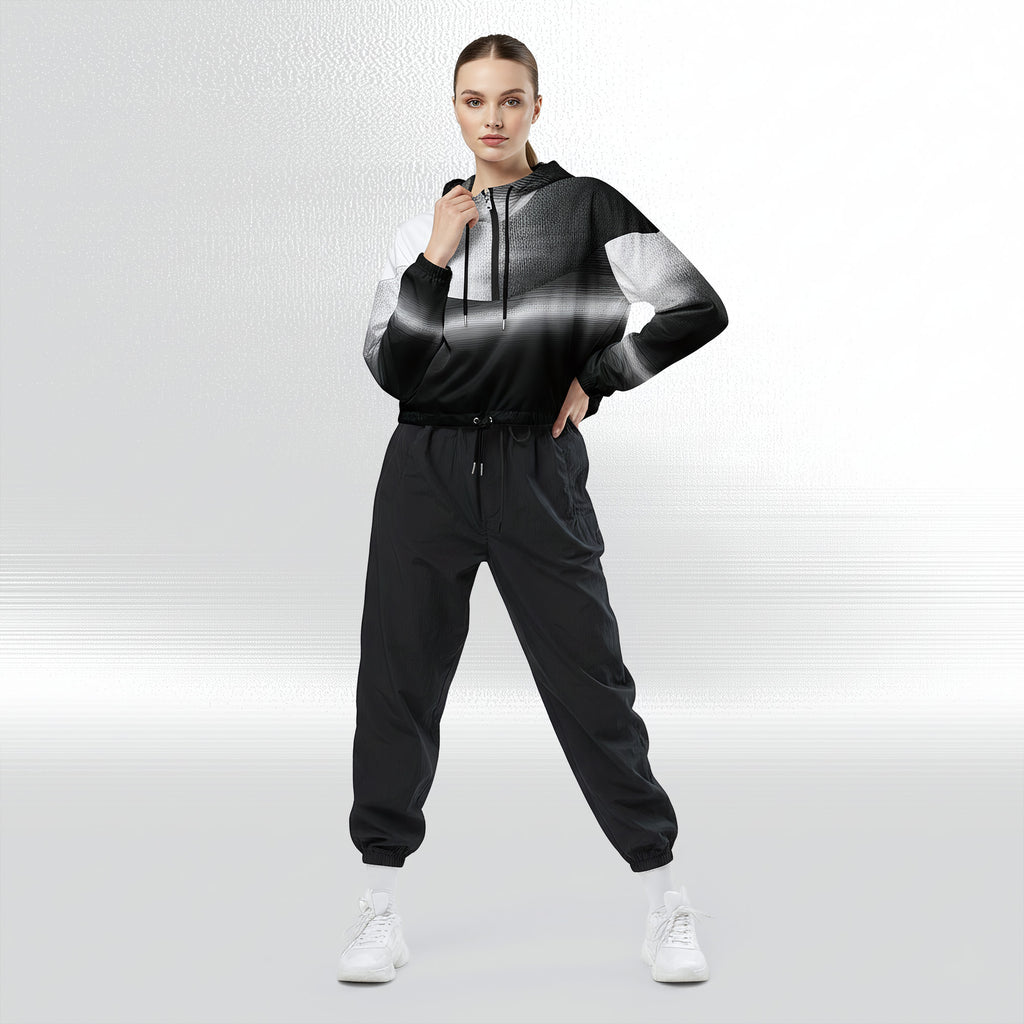 Full‑body view of a model standing in the HER Cropped Dither Distortion Windbreaker. The hooded jacket, with its abstract black‑to‑white gradient print and half‑zip closure, is worn with black trousers and white shoes. The scene has a minimalist grey background