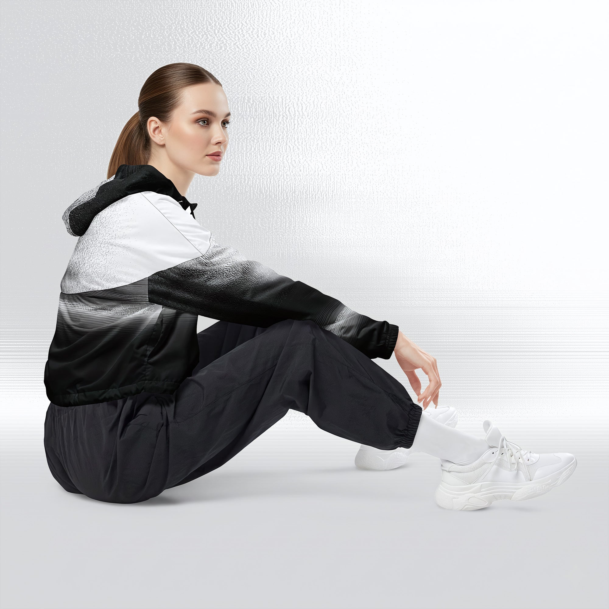 Side profile of a woman seated cross‑legged in the HER Cropped Dither Distortion Windbreaker. The cropped jacket shows a digital dither gradient from white to black, with the hood lowered and a drawcord at the hem. She wears black cargo trousers and white trainers, photographed against a neutral grey backdrop
