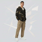 Front portrait of a male model wearing the Form Finding Design men’s Shadow Drip Hoodie (No.10), zipped up and hands by his sides; the hoodie’s abstract tan and olive print contrasts with his khaki cargo trousers, set against a pale grey background with faint curved motifs.