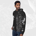 A man wearing a black pullover hoodie with a detailed black and white graphic print on the front and sleeves, paired with blue jeans, set against a light gray patterned background.