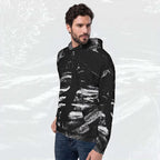 A man wearing a black pullover hoodie with a white abstract brushstroke pattern, paired with blue jeans, set against a light gray textured background.