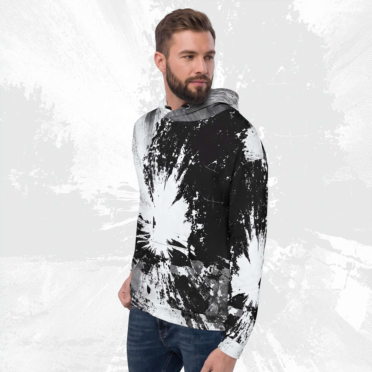 A man wearing a black and white abstract paint splatter hoodie with a gray hood lining, paired with blue jeans, set against a textured light gray background.