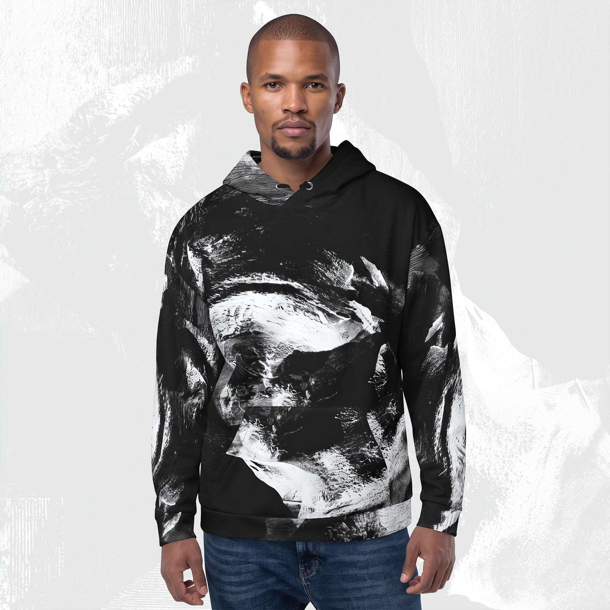 A man wearing a black pullover hoodie with a large white and gray abstract brushstroke print, paired with blue jeans, set against a light gray textured background.