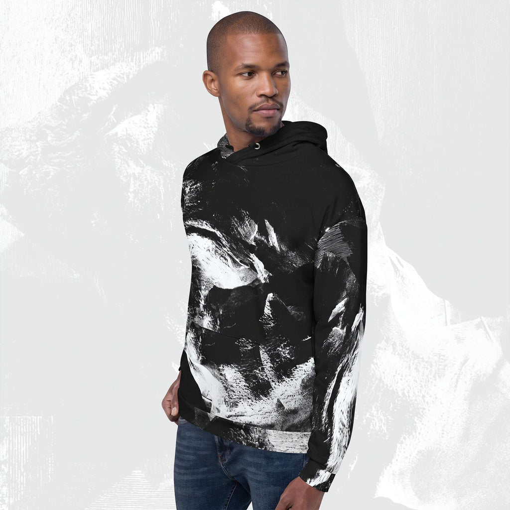 A man wearing a black pullover hoodie with a bold white abstract brushstroke pattern, paired with blue jeans, set against a light gray textured background.