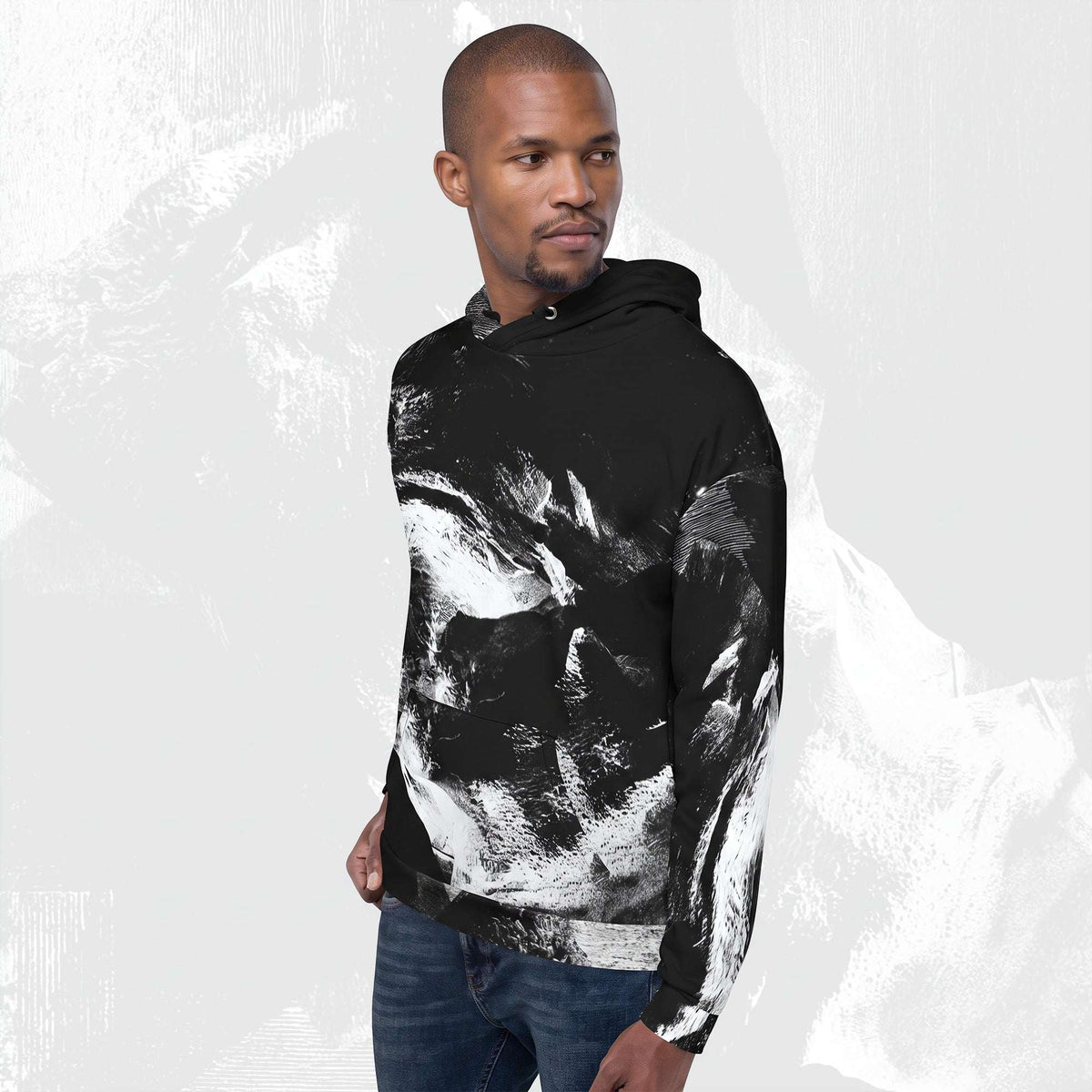 A man wearing a black pullover hoodie with a bold white abstract brushstroke pattern, paired with blue jeans, set against a light gray textured background.