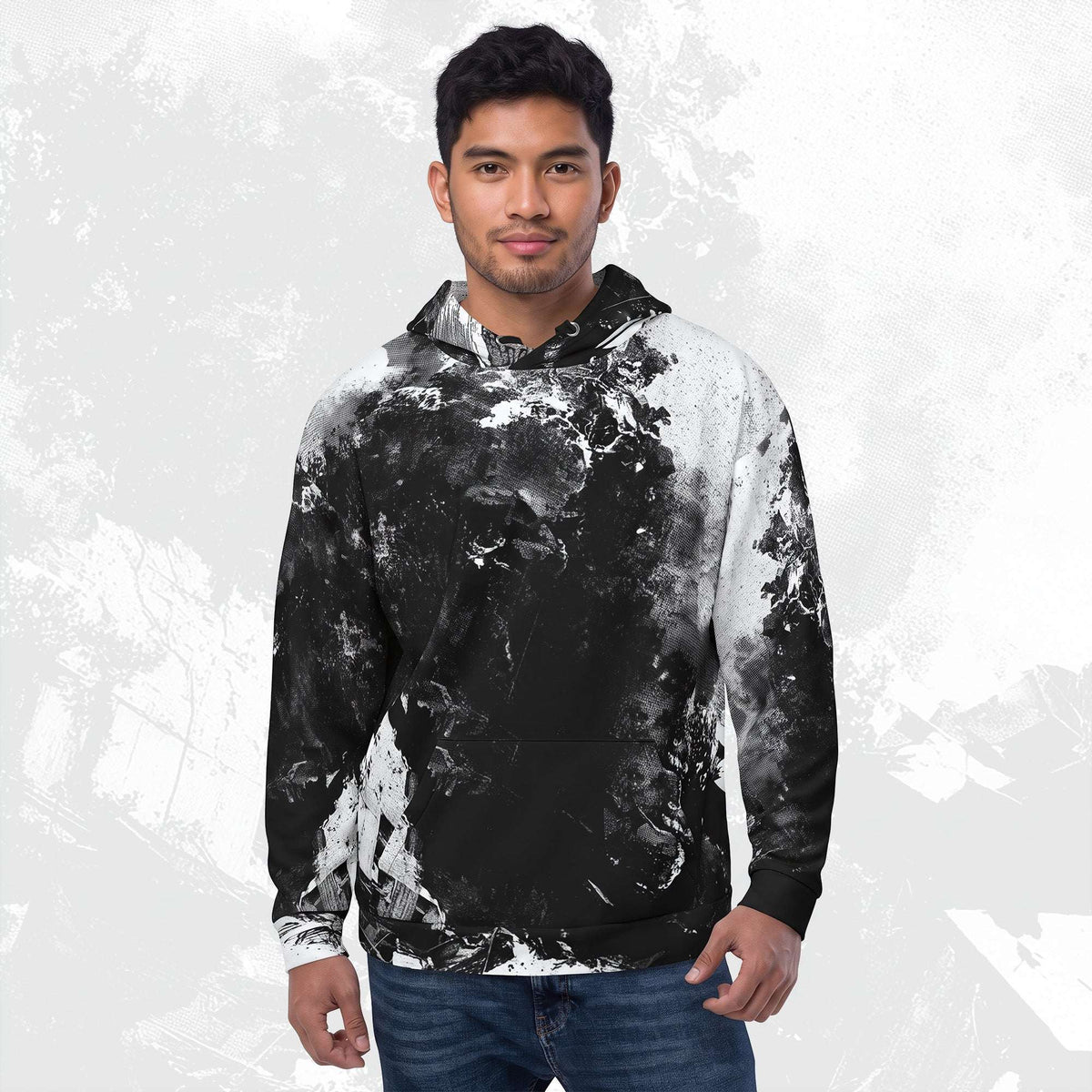 A man wearing a black and white marbled hoodie with blue jeans, standing against a textured light gray background.