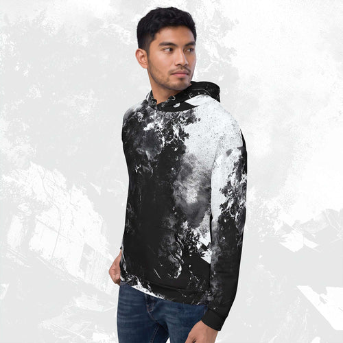 A man wearing a black and white abstract splatter print hoodie with blue jeans, standing against a light gray textured background.