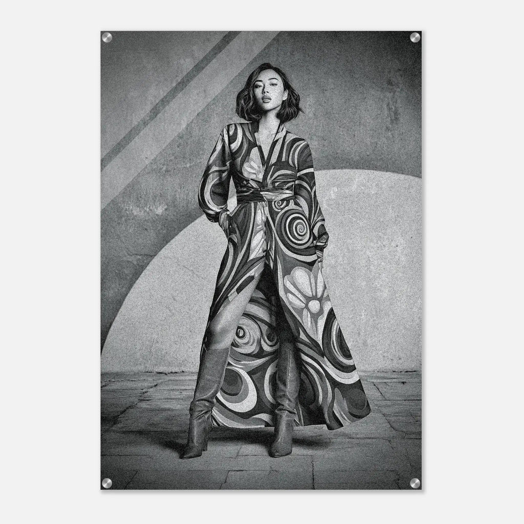 A black and white fashion poster featuring a woman wearing a long, flowing patterned coat and tall boots, standing against a textured geometric wall. The poster is shown hanging on a light gray wall with visible metal mounting hardware in the corners.