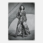 A black and white fashion poster featuring a woman wearing a long, flowing patterned coat and tall boots, standing against a textured geometric wall. The poster is shown hanging on a light gray wall with visible metal mounting hardware in the corners.
