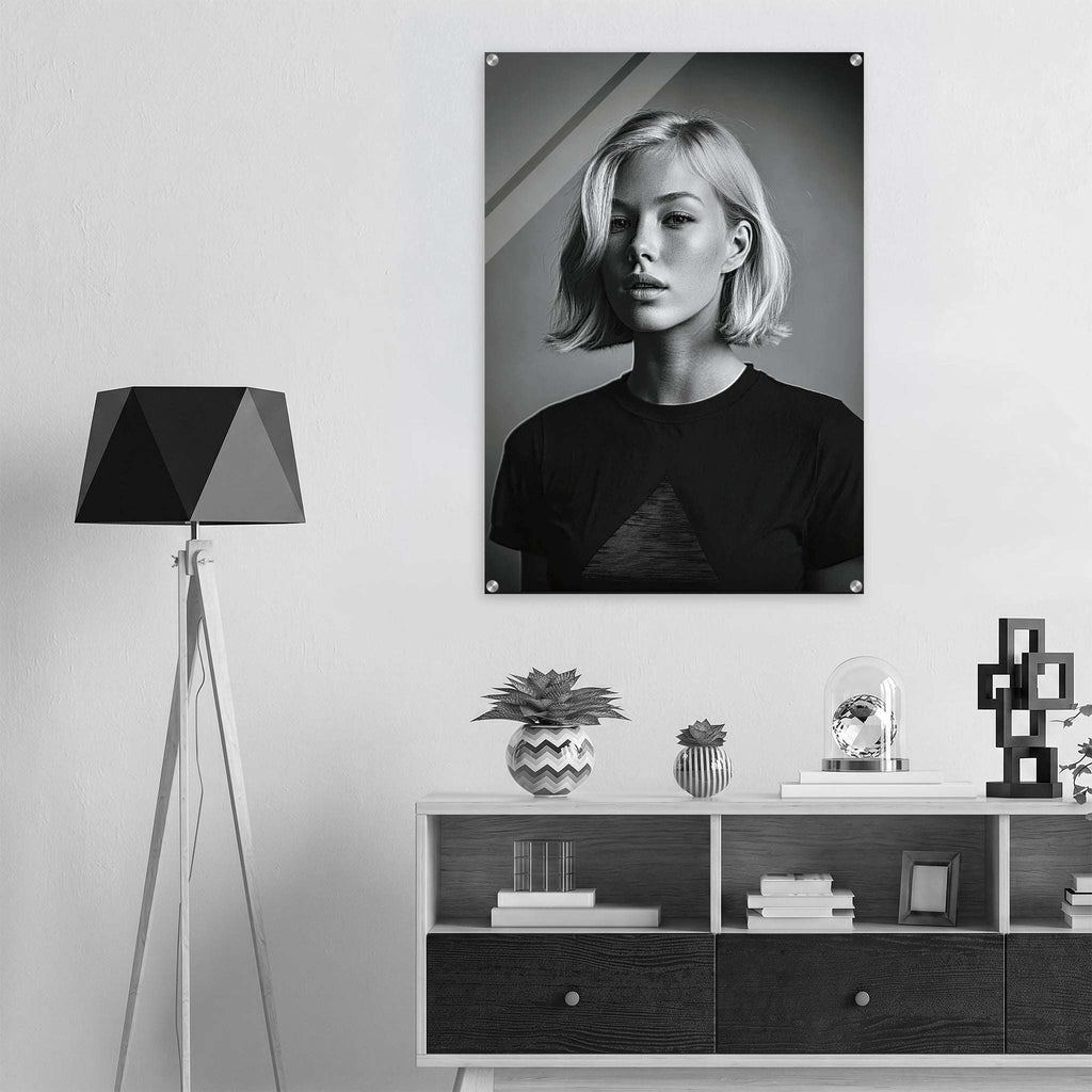 A black and white portrait poster of a woman with short blonde hair, displayed on a light gray wall above a modern white and dark wood console shelf. The room is styled with a black geometric tripod floor lamp on the left and decorative objects on the shelf.
