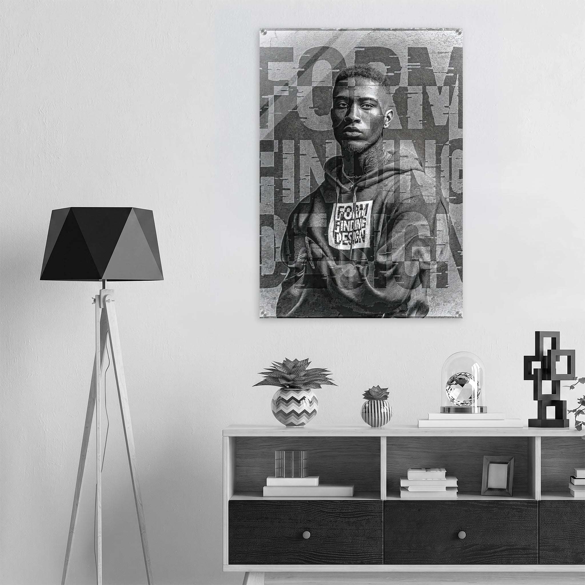 A black and white poster print of a young man wearing a hoodie with the text 'FORM FINDING DESIGN' displayed on a wall above a modern white and dark wood console shelf, next to a black tripod floor lamp.