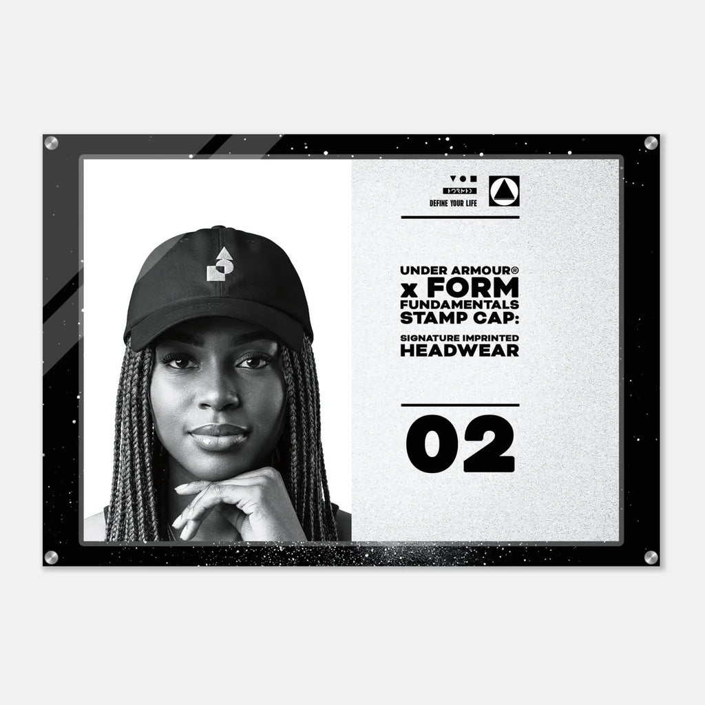 A black-framed acrylic wall art print featuring a black-and-white portrait of a woman wearing a cap on the left, with text on the right reading 'UNDER ARMOUR x FORM FUNDAMENTALS STAMP CAP' and '02'.