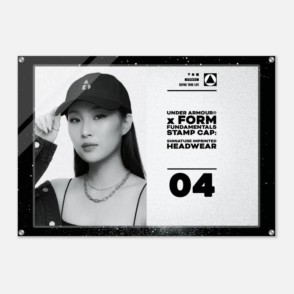 A black-framed acrylic wall plaque featuring a black-and-white portrait of a woman wearing a baseball cap, alongside text reading 'UNDER ARMOUR x FORM FUNDAMENTALS STAMP CAP' and 'SIGNATURE IMPRINTED HEADWEAR' with the number 04.
