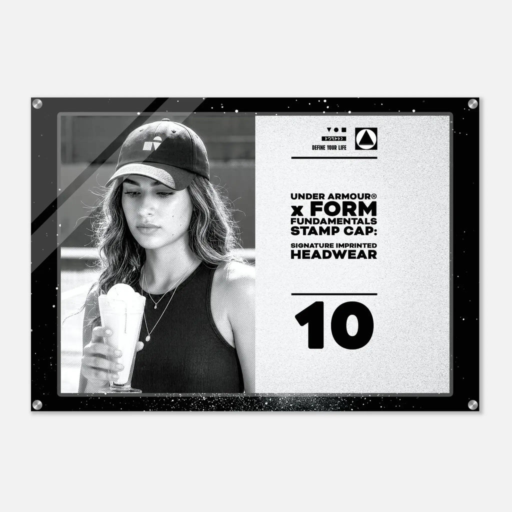 A rectangular black-framed acrylic or glass wall print featuring a black-and-white photo of a woman wearing a baseball cap and holding an iced drink, alongside printed text that reads 'UNDER ARMOUR x FORM FUNDAMENTALS STAMP CAP: SIGNATURE IMPRINTED HEADWEAR' and the number '10'.