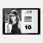 A rectangular black-framed acrylic or glass wall print featuring a black-and-white photo of a woman wearing a baseball cap and holding an iced drink, alongside printed text that reads 'UNDER ARMOUR x FORM FUNDAMENTALS STAMP CAP: SIGNATURE IMPRINTED HEADWEAR' and the number '10'.