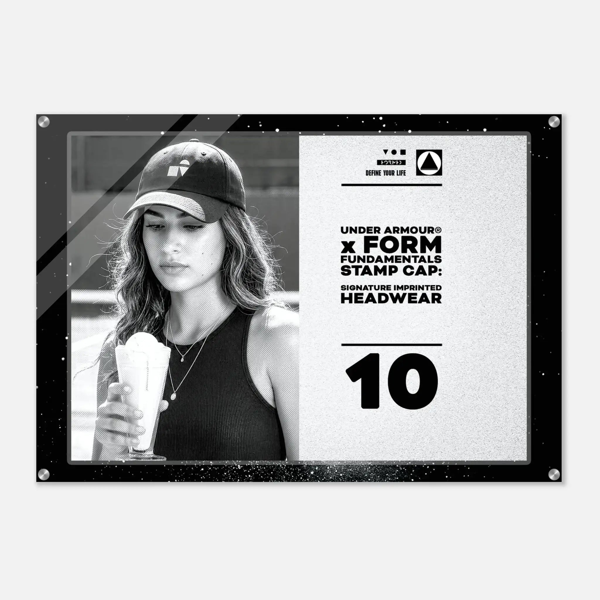 A rectangular black-framed acrylic or glass wall print featuring a black-and-white photo of a woman wearing a baseball cap and holding an iced drink, alongside printed text that reads 'UNDER ARMOUR x FORM FUNDAMENTALS STAMP CAP: SIGNATURE IMPRINTED HEADWEAR' and the number '10'.