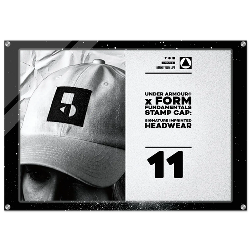 A framed black-and-white promotional print featuring a close-up of a person wearing a baseball cap with a large Under Armour stamp logo. The right side includes text reading 'UNDER ARMOUR x FORM FUNDAMENTAL STAMP CAP: SIGNATURE IMPRINTED HEADWEAR' and the number 11.