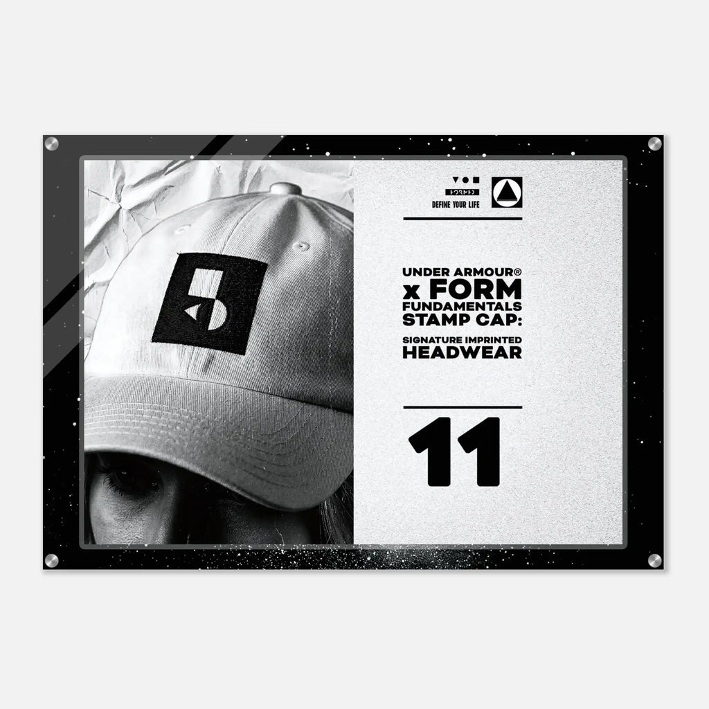 A rectangular black-framed print or poster on a light gray background. The artwork shows a black-and-white close-up of a baseball cap with a square Under Armour logo patch, alongside white text reading 'UNDER ARMOUR x FORM FUNDAMENTALS STAMP CAP' and the number '11'.