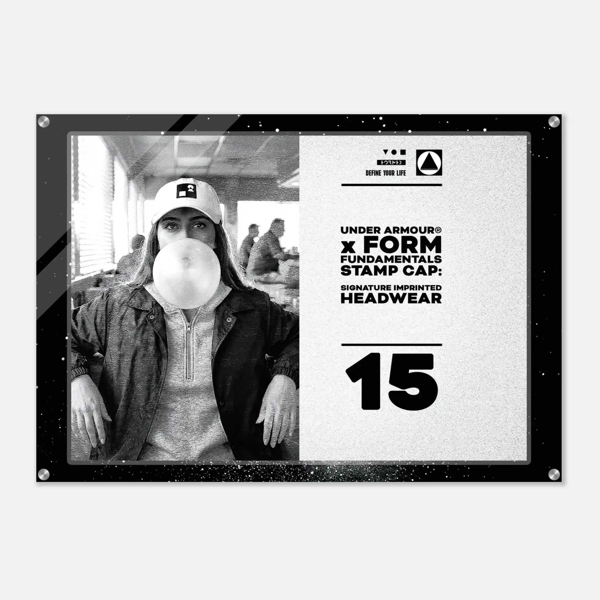 A rectangular acrylic wall art print with rounded corners and visible mounting hardware, featuring a black-and-white photo of a person wearing a cap and blowing a bubble gum bubble, alongside white text reading 'UNDER ARMOUR x FORM FUNDAMENTALS STAMP CAP: SIGNATURE IMPRINTED HEADWEAR' and the number '15'.