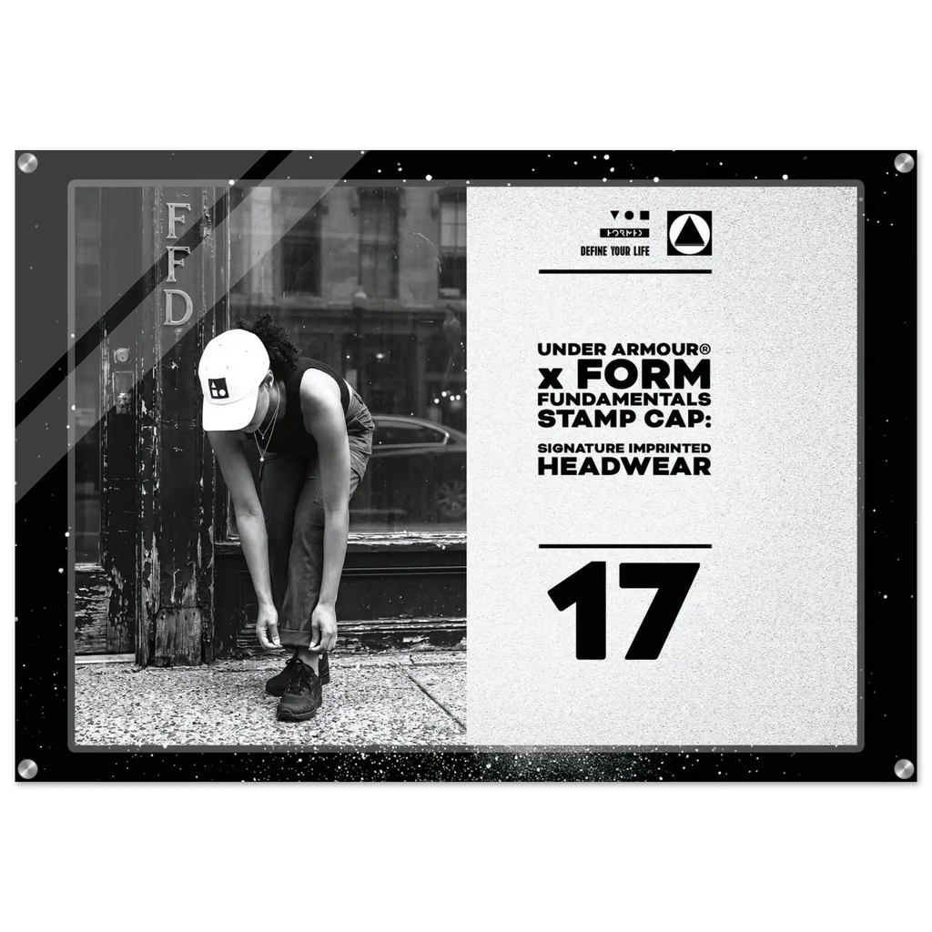 A rectangular framed stamp cap print featuring a black-and-white street photo of a person tying shoes beside a pole, alongside large text reading 'UNDER ARMOUR x FORM FUNDAMENTALS STAMP CAP: SIGNATURE IMPRINTED HEADWEAR' and the number '17'.
