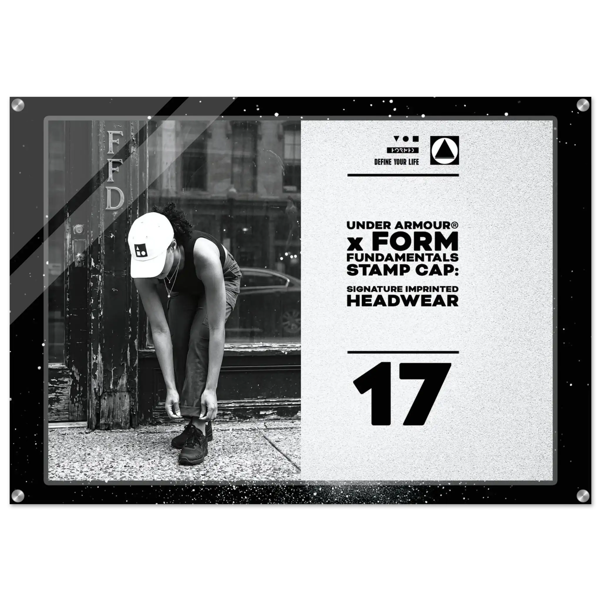 A rectangular framed stamp cap print featuring a black-and-white street photo of a person tying shoes beside a pole, alongside large text reading 'UNDER ARMOUR x FORM FUNDAMENTALS STAMP CAP: SIGNATURE IMPRINTED HEADWEAR' and the number '17'.