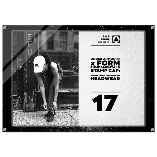A rectangular framed stamp cap print featuring a black-and-white street photo of a person tying shoes beside a pole, alongside large text reading 'UNDER ARMOUR x FORM FUNDAMENTALS STAMP CAP: SIGNATURE IMPRINTED HEADWEAR' and the number '17'.