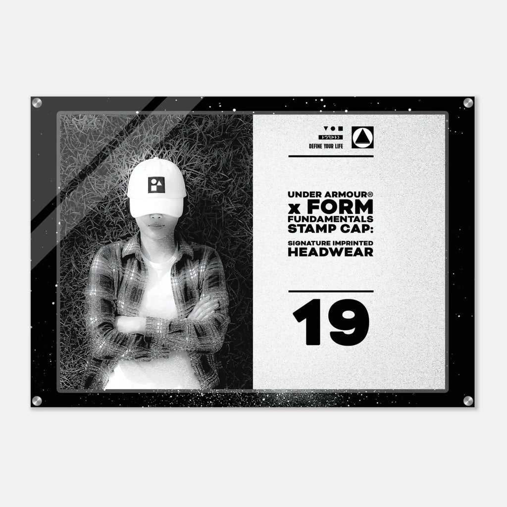 A rectangular acrylic wall art print with a black frame and corner screws, featuring a black-and-white split design. The left side shows a grayscale photo of a person wearing a white cap, and the right side has text reading 'UNDER ARMOUR x FORM FUNDAMENTALS STAMP CAP SIGNATURE IMPRINTED HEADWEAR' with the number 19.