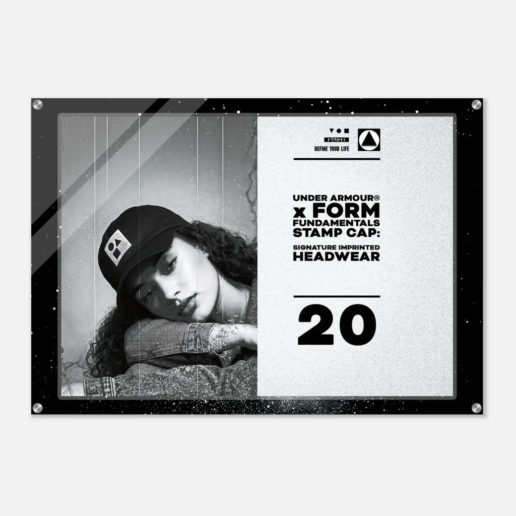 A rectangular black-framed acrylic wall sign featuring a black-and-white portrait of a woman wearing a cap on the left and printed text on the right, including 'UNDER ARMOUR' and 'FORM FUNDAMENTALS STAMP CAP SIGNATURE IMPERFECT HEADWEAR' with the number 20, shown against a light gray background.